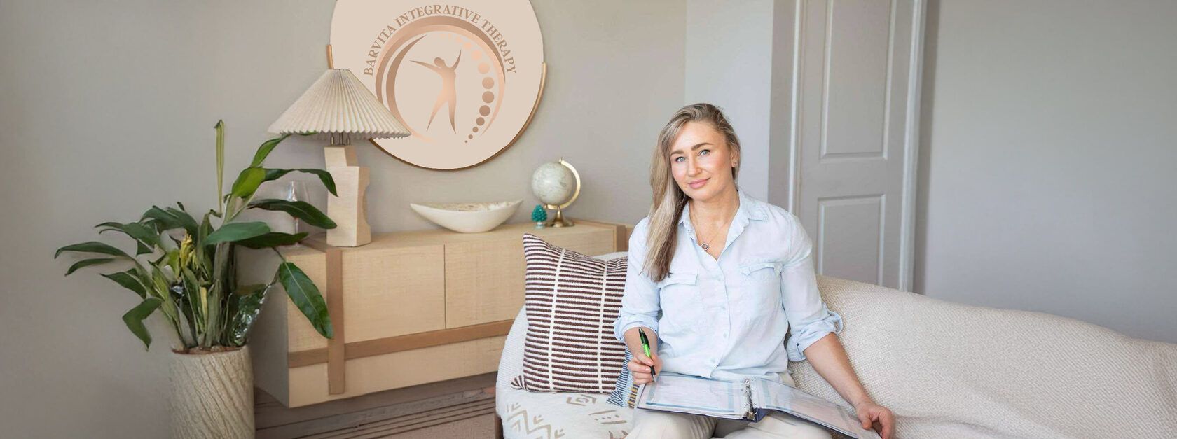 Violeta Barbolyuk in a light blue shirt, seated on a white sofa with a folder and a pen, under a round Barvita Integrative Therapy sign